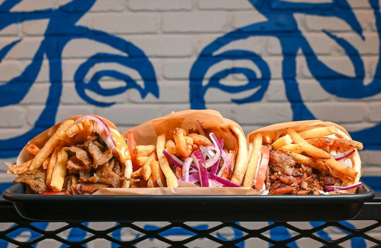 Three Greek gyros topped with fries and onions in front of THE GREEK mural